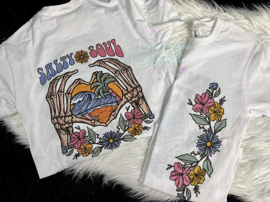 White tee that has ocean on back with salty soul on it and flowers on the front.